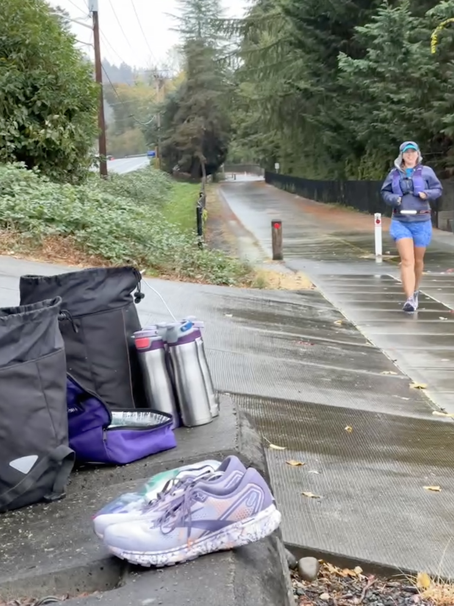 A picture of Dana Lewis running down the rainy paved trail, with resupply gear (dry shoes, water, fuel) in the foreground of the picture. She's wearing shorts, a rain jacket, and a rain hat. She is smiling and around 12 miles into her eventual 82 mile run).