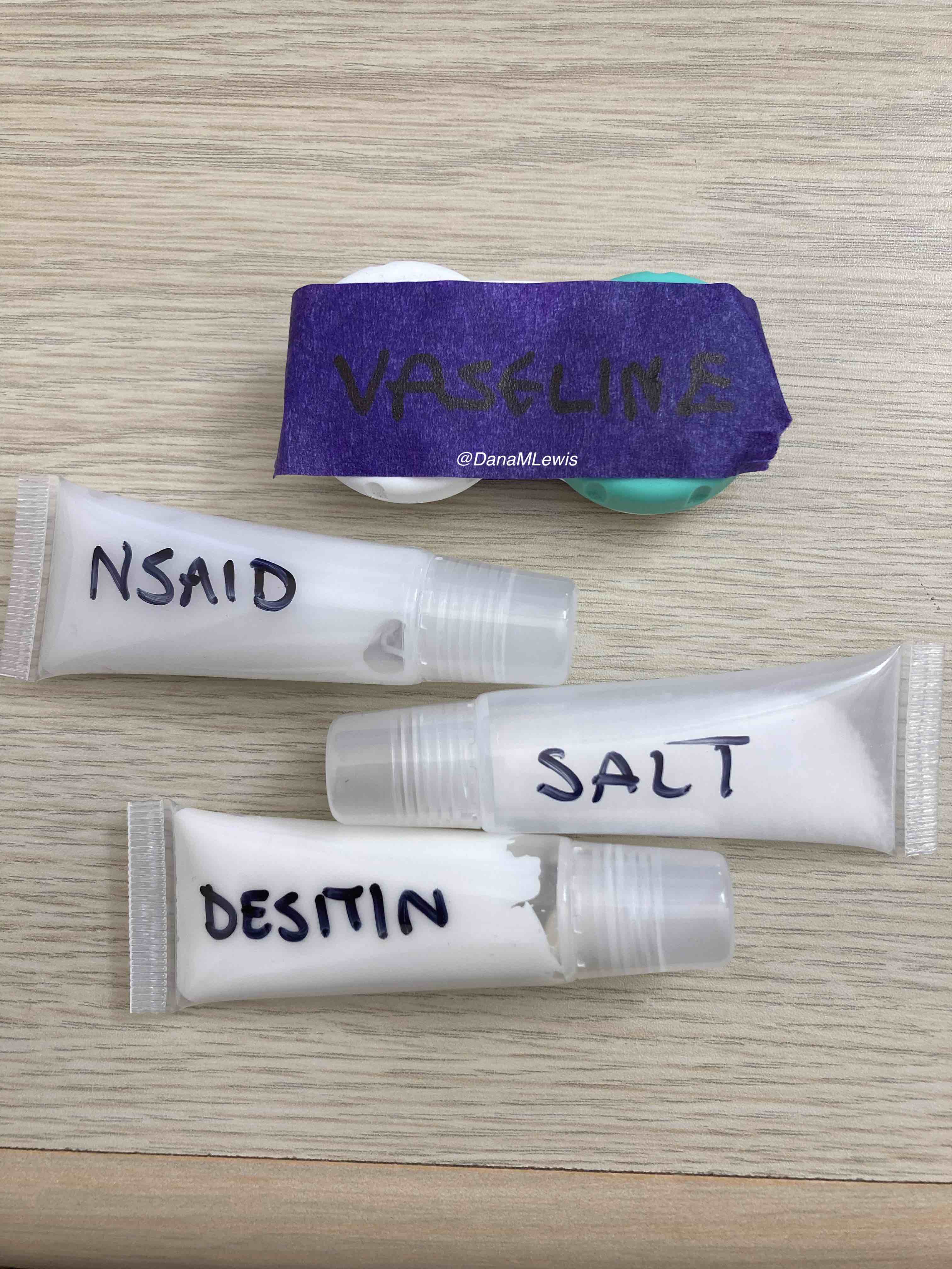A contact case with a strip of purple painter tape that has "vaseline" written on it in sharpie; and three lip gloss tubes filled and marked with "NSAID", "salt", and "desitin". 
