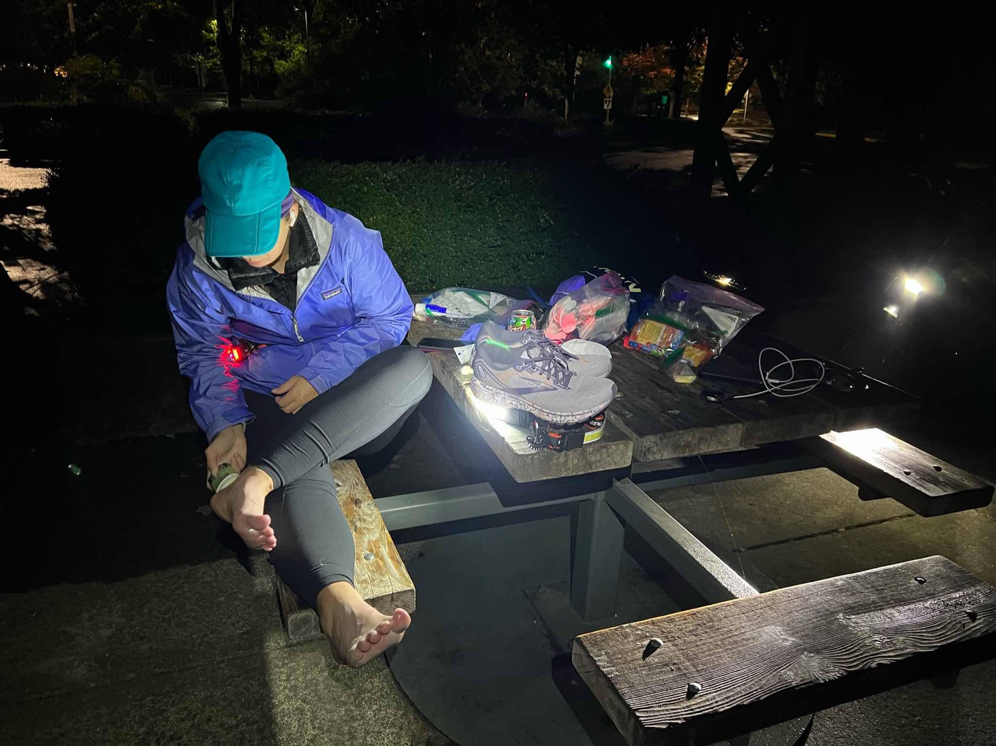 Dana sits on a bench at a picnic table in a public park. It is dark. She is wearing long rain pants, a rain jacket, and a rain hat and is bent over her bare feet, applying lubrication. It is dark and nighttime, so she has an extra waistlamp on the table illuminating her feet. Other ultrarunning supplies are strewn across the table.