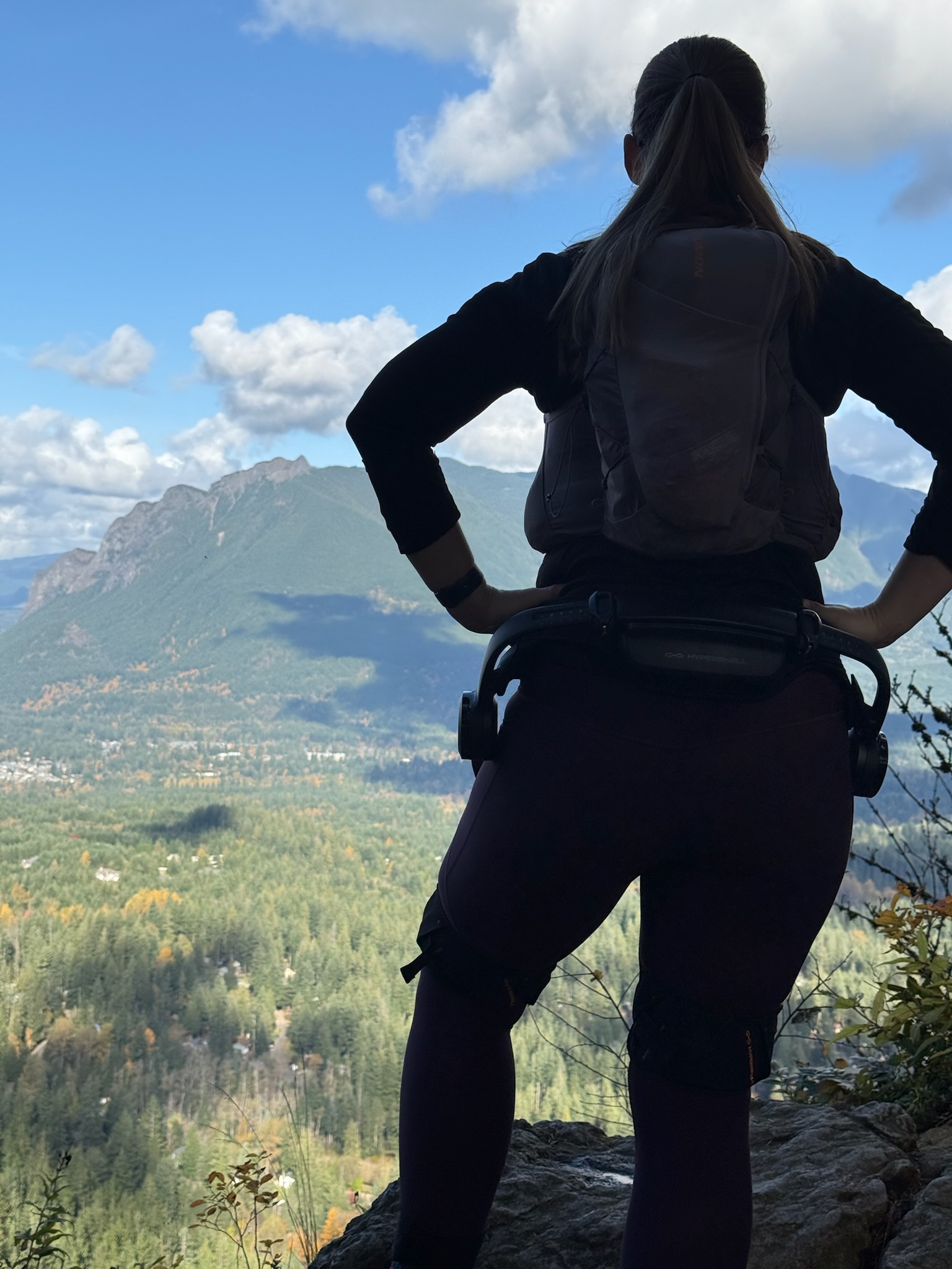 Dana is a dark silhouette where you can see the sides of the exoskeleton's motors next to each hip, looking down at the sunlit valley with Rattlesnake Lake and Ledge ahead of her