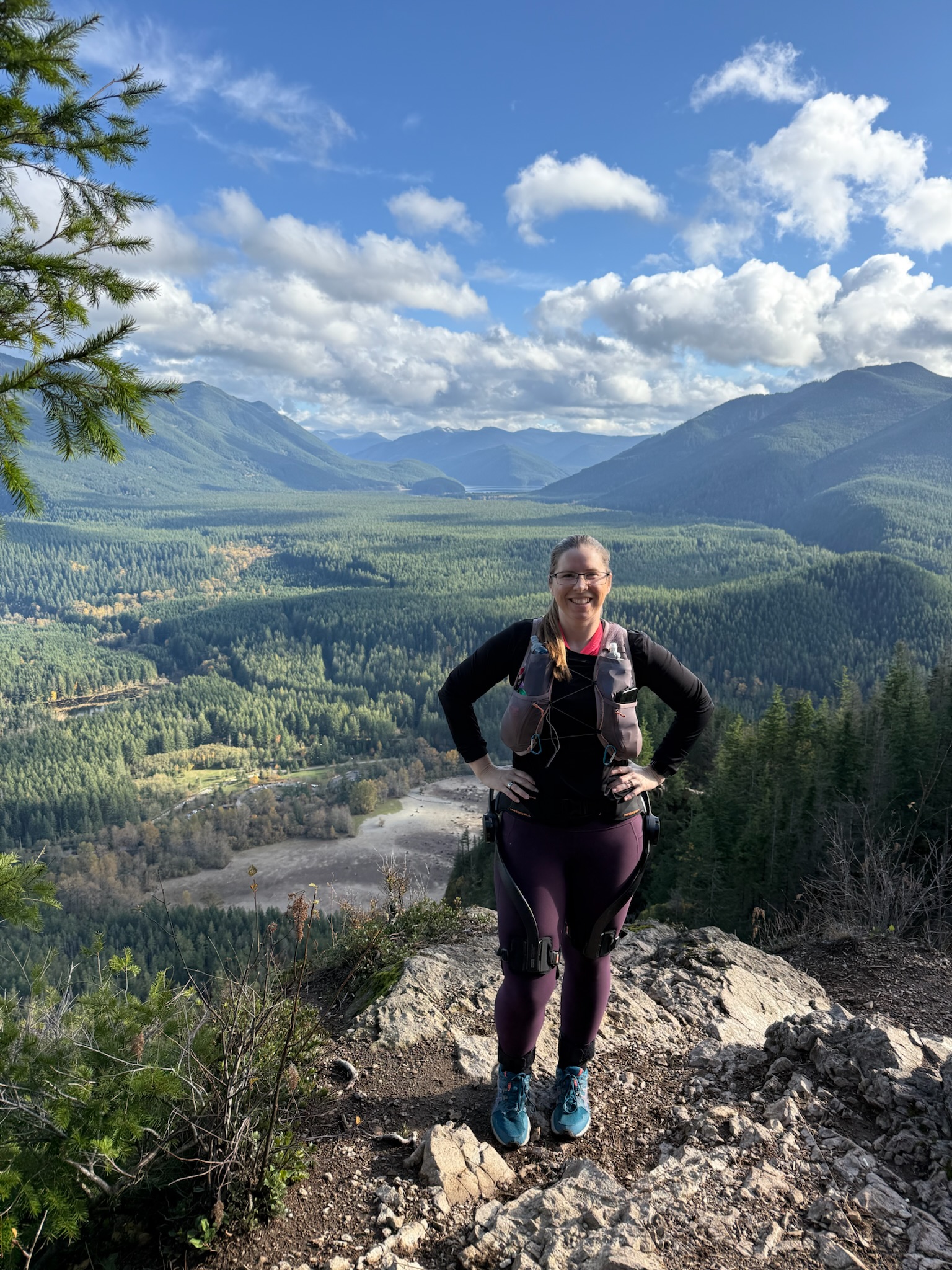Dana is facing the camera, standing not very near the edge of the second ledge, with views of the dry Rattlesnake Lake and Rattelsnake Ledge behind her. It is a bright sunny day with the valley lit behind her.